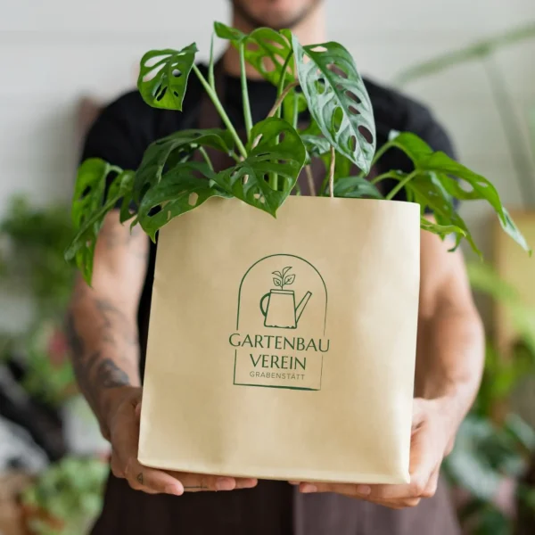 Gartenbauverein Grabenstätt Brand Design auf Papiertüte mit Monstera Pflanze von einem Mann gehalten